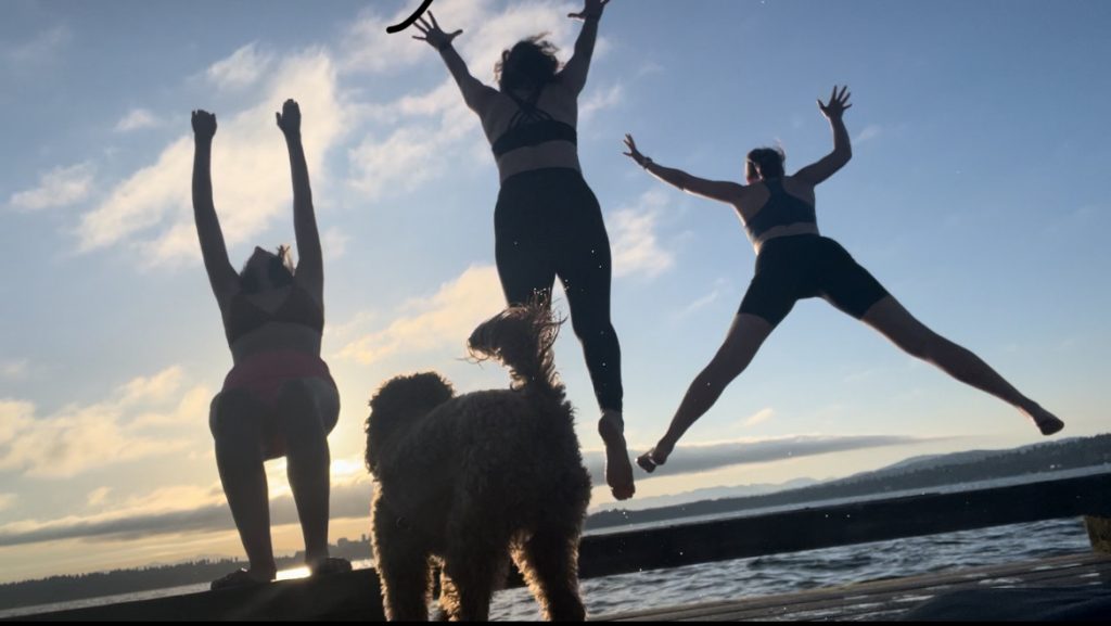 silhouette of 3 individuals jumping into a lake, arms extended, with a cute puppy in the foreground