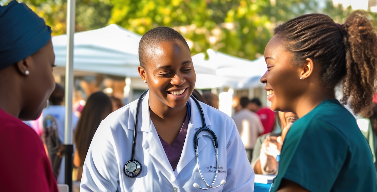 Smiling women interact at an outdoor community health event with tents and other attendees in the background.